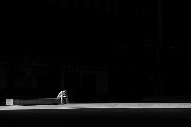 Person sitting alone on a bench in a dark, empty space, illuminated by a spotlight, representing solitude and contemplation.