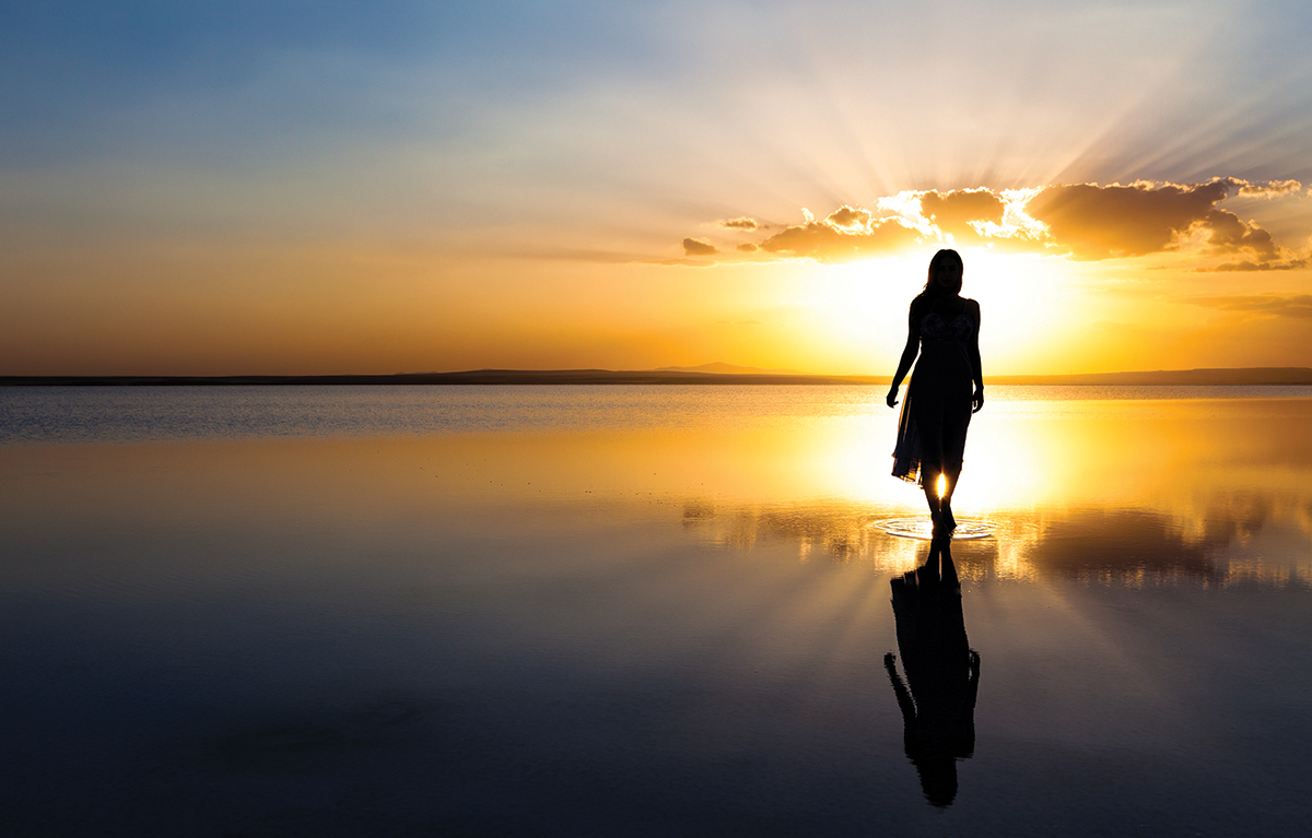 Silhouette of a woman walking on water reflecting a stunning sunset sky, creating a tranquil and serene scene.