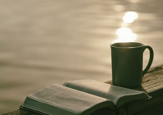 Open book and mug on a wooden deck by a lake, reflecting sunlight for a serene relaxation moment.