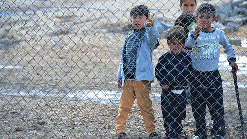 Children standing behind a chain-link fence making peace signs, conveying hope and resilience.