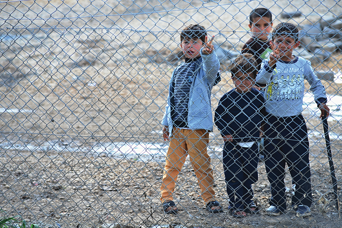 Children standing behind a chain-link fence making peace signs, conveying hope and resilience.