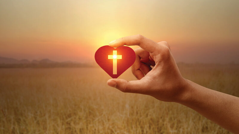 Hand holding a heart with a cross against a sunset, symbolizing faith and love.