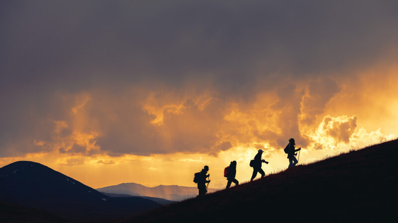 Silhouettes of hikers climbing a hill at sunset, with dramatic clouds and mountains in the background.