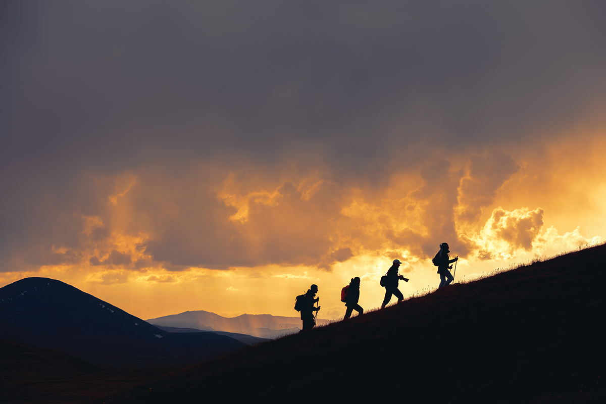 Silhouettes of hikers climbing a hill at sunset, with dramatic clouds and mountains in the background.