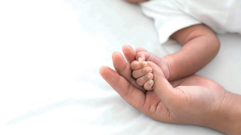 Baby holding parent's hand, symbolizing love and connection. Close-up on soft bedsheet background.