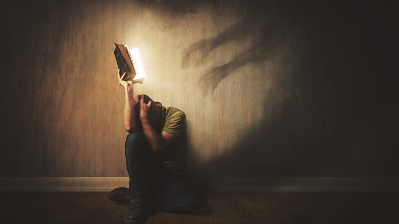 Person shielding with glowing book from shadowy hands on wall, symbolizing fear and protection.