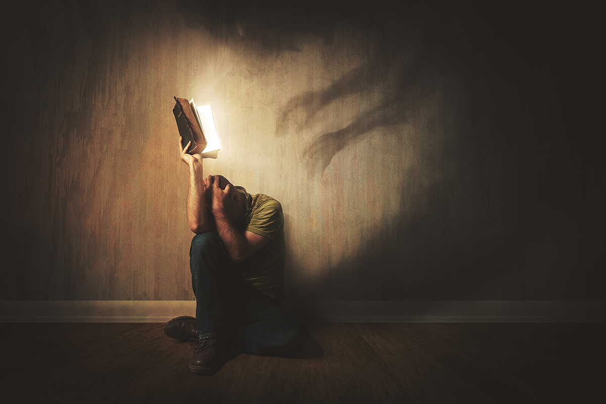 Person shielding with glowing book from shadowy hands on wall, symbolizing fear and protection.