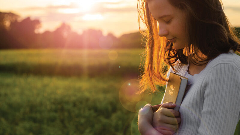 Young woman holding a Bible, praying peacefully outdoors at sunset, with a serene expression.
