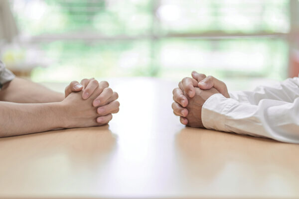 Two individuals with clasped hands sitting opposite each other at a table, suggesting a conversation or negotiation.