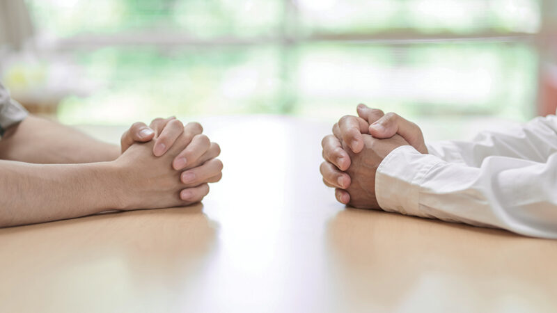 Two individuals with clasped hands sitting opposite each other at a table, suggesting a conversation or negotiation.