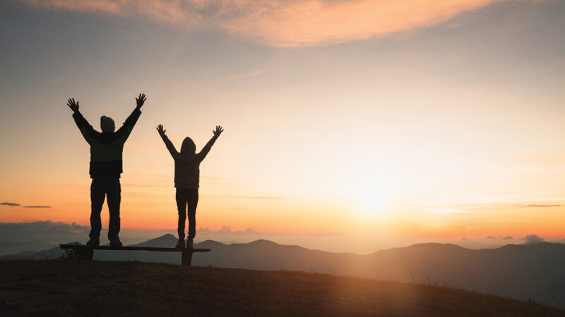 Two people celebrating sunrise on a mountain with arms raised, capturing a moment of joy and freedom.
