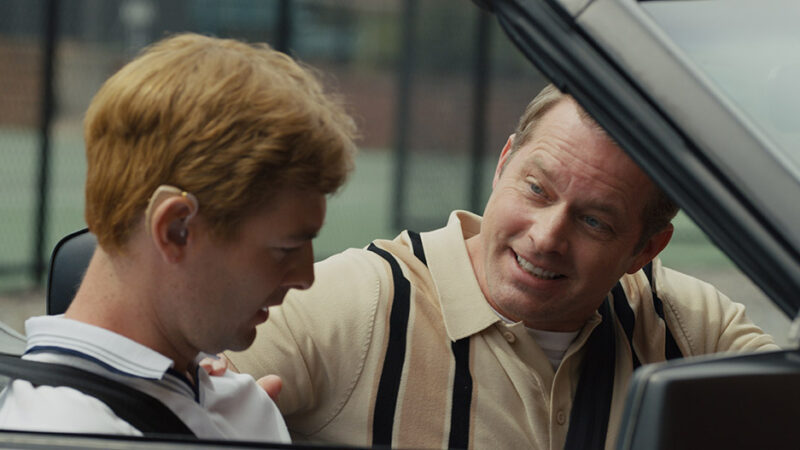 Man smiling at another man in a car with a tennis backdrop.