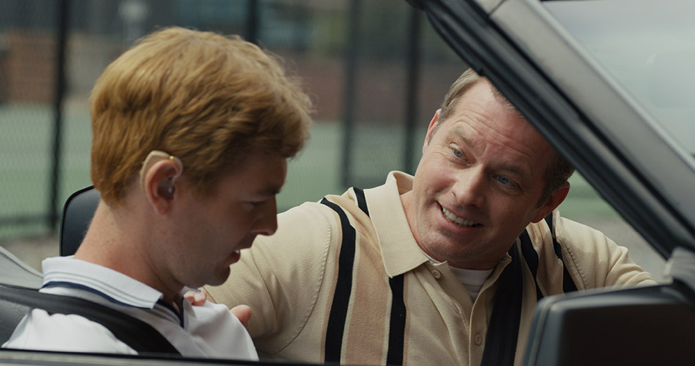 Man smiling at another man in a car with a tennis backdrop.
