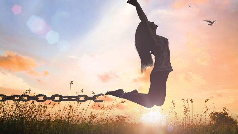 Silhouette of woman breaking chains, leaping in freedom at sunset with birds in sky.