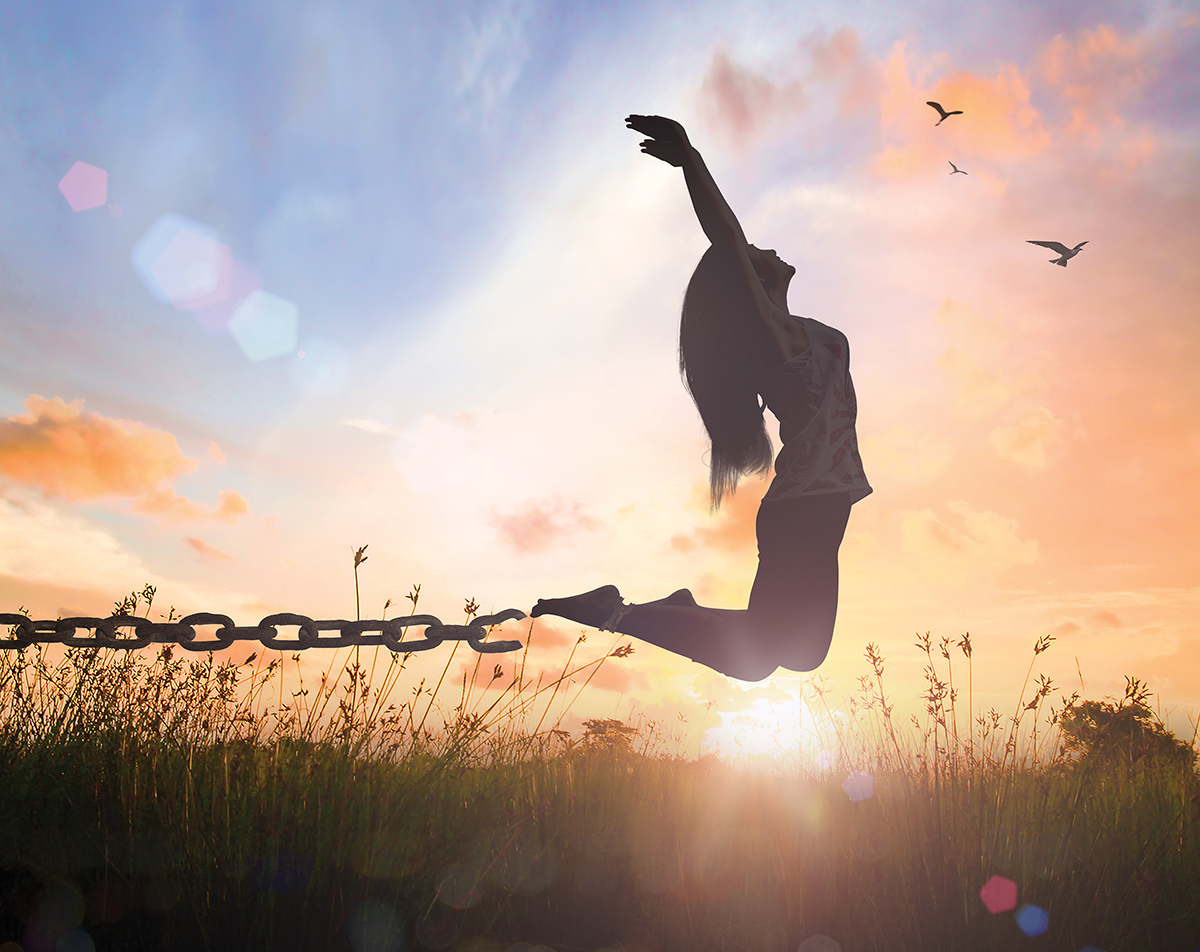 Silhouette of woman breaking chains, leaping in freedom at sunset with birds in sky.