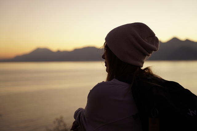 Woman in beanie gazes at serene sunset over lake, mountains in the distance. Tranquil outdoor moment.