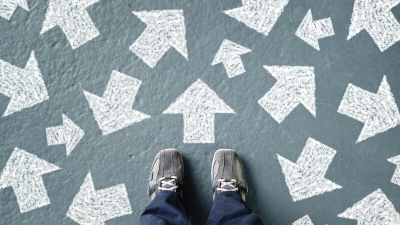 Feet standing on pavement with many chalk-drawn arrows pointing in multiple directions, symbolizing decision-making or choices.