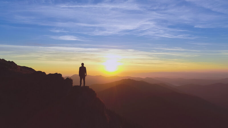 Person standing on a mountain peak during sunrise, overlooking hills and a colorful sky.