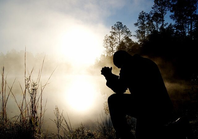 Silhouette of a person kneeling in prayer by a misty lake at sunrise, surrounded by trees and grass.