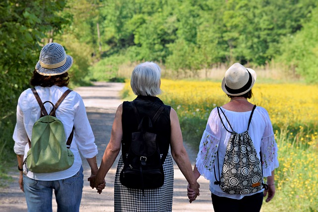 Three women walking hand in hand on a scenic path surrounded by nature, carrying backpacks and wearing hats.