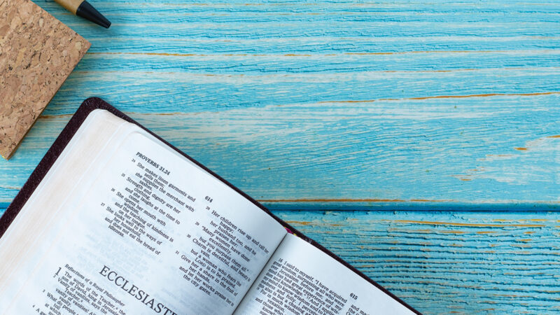Open Bible on Ecclesiastes with cork notebook and pen on blue wooden background.