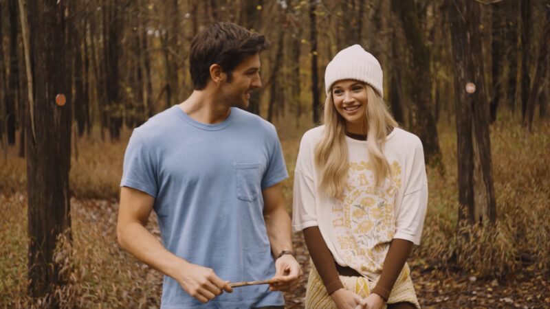 Couple smiling and walking in a forest during autumn, wearing casual clothing and enjoying nature.