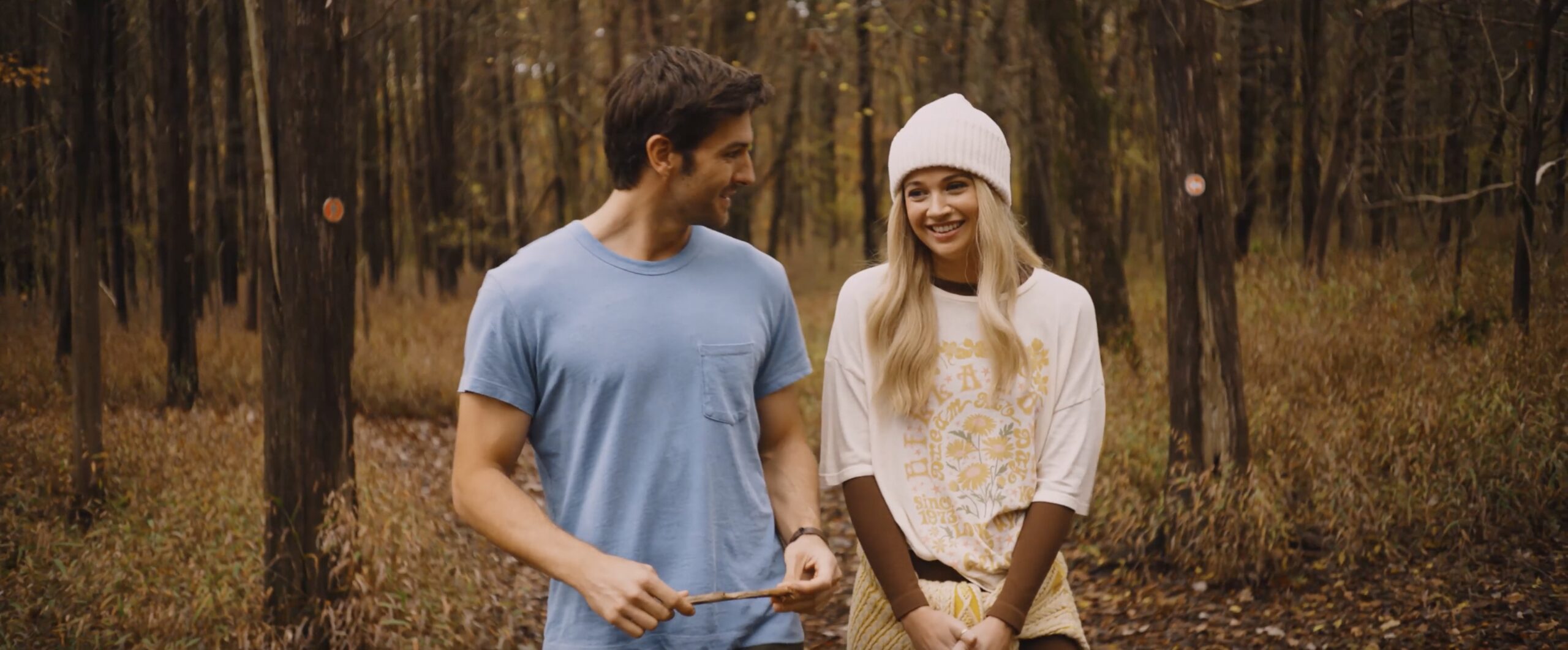 Couple smiling and walking in a forest during autumn, wearing casual clothing and enjoying nature.