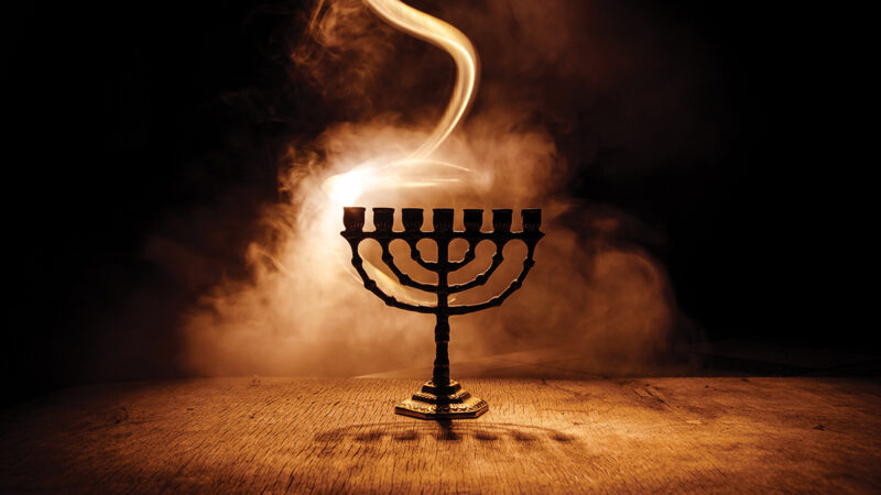 Menorah with nine branches casting shadow in smoky, dimly lit atmosphere, symbolizing a cultural celebration.