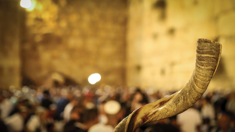 Shofar horn in focus against a blurred background of a stone wall and crowd at a religious gathering.