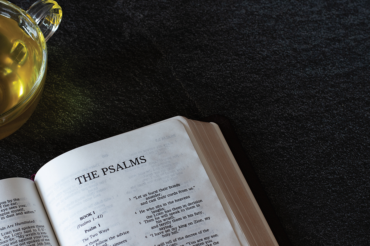 Open Bible showing the Book of Psalms next to a cup of tea on a dark surface.