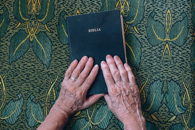 Elderly hands holding a Bible on green patterned cloth, symbolizing faith and spirituality.