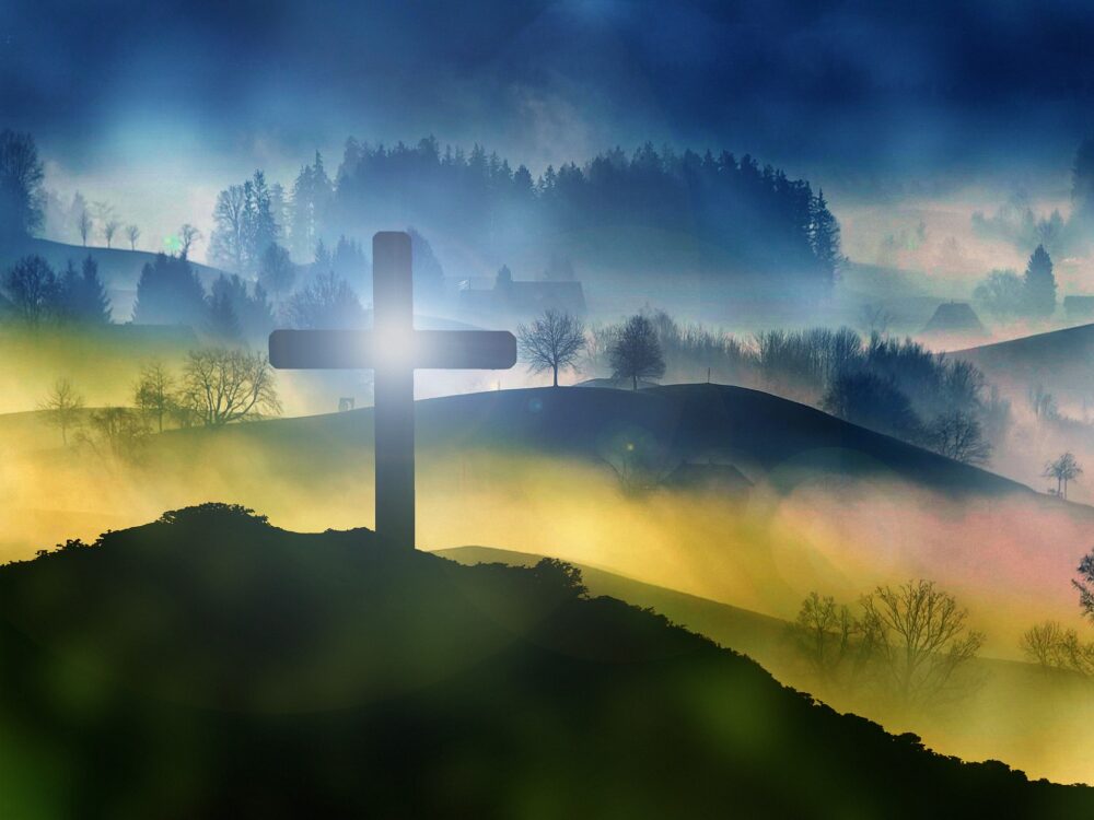 Silhouette of a cross on a hill with a misty, forested landscape in the background and a bright light shining through the cross at sunrise or sunset.