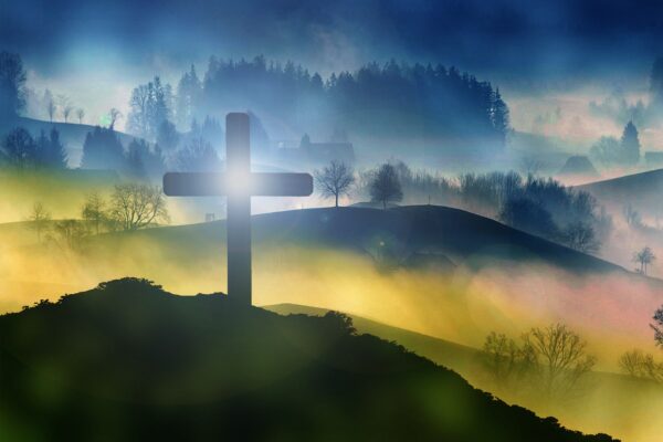 Silhouette of a cross on a misty hill with sunrise, forest, and foggy landscape in the background.