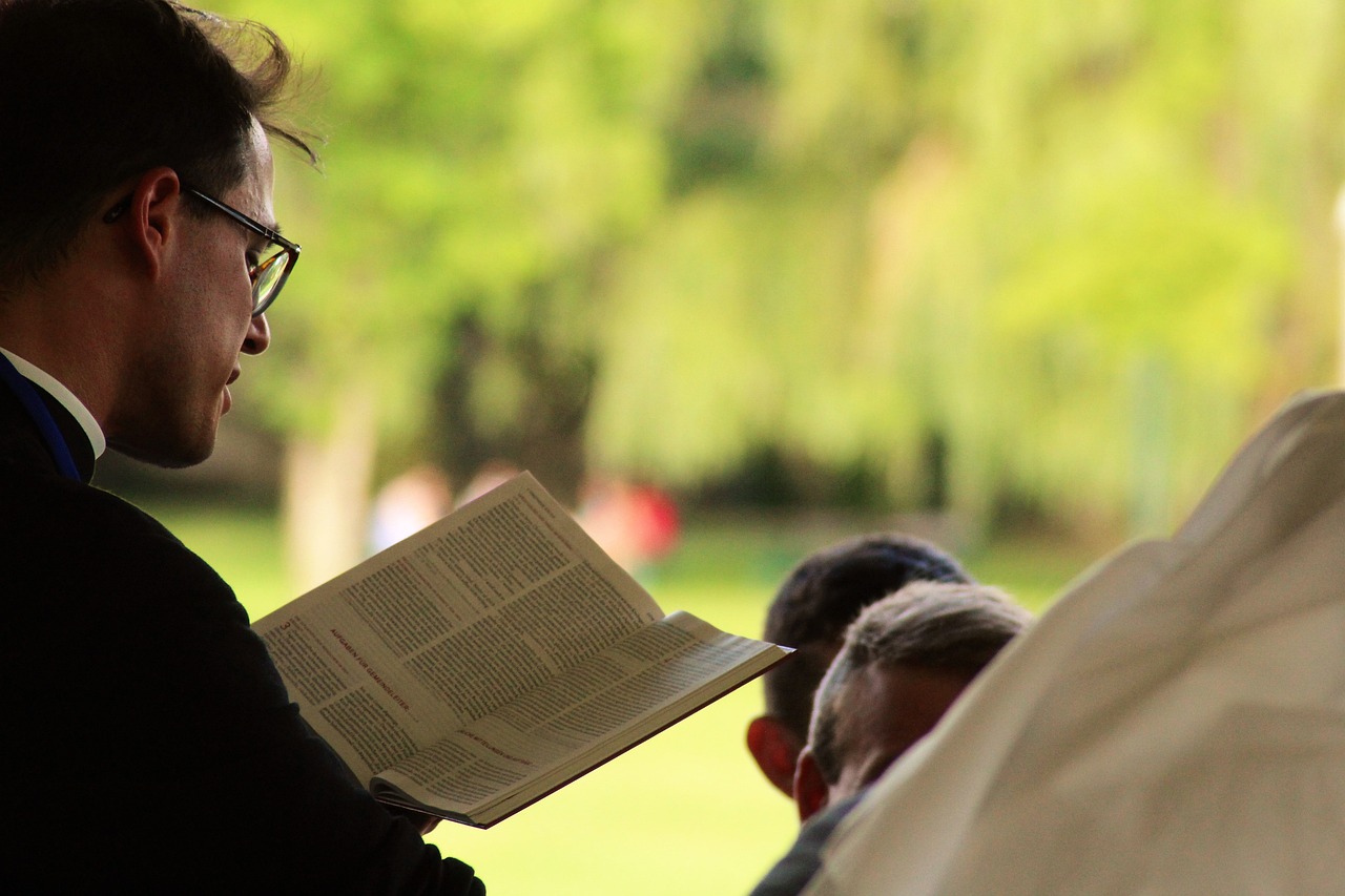 Man reading a book aloud outdoors, surrounded by listeners, set against a blurred green background.