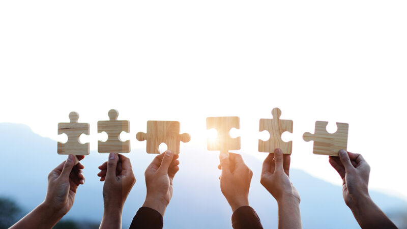 Teamwork and partnership concept. Hands are raised against a bright, white sky, each holding a piece of a wooden jigsaw puzzle. teamwork, problem solving, Charity, volunteer. Unity, team business.