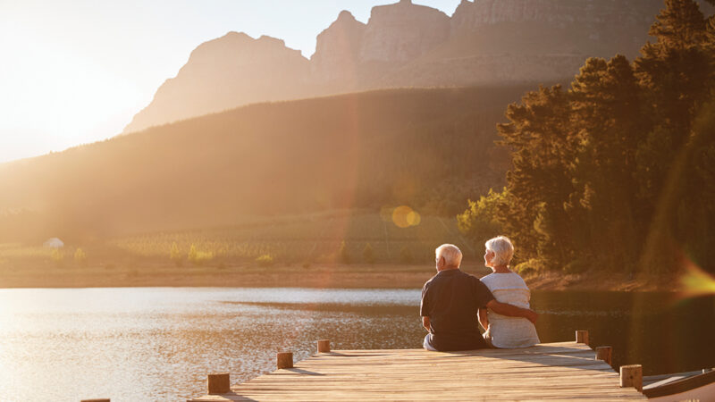 Romantic Senior Couple Sitting On Wooden Jetty By Lake