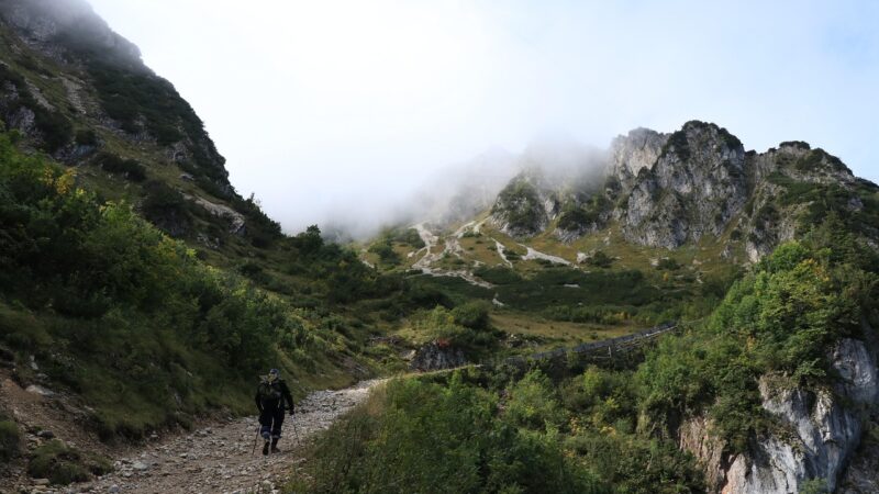 Hiker on a mountain trail surrounded by lush greenery and misty peaks, creating a serene and adventurous landscape.