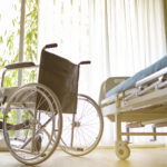 A bright hospital room featuring a wheelchair beside a medical bed, surrounded by sunlight and greenery.