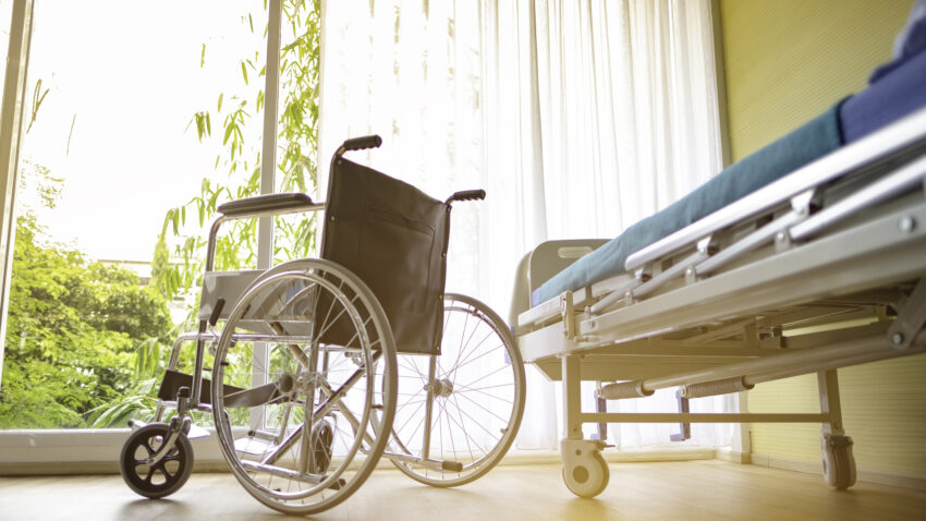 A bright hospital room featuring a wheelchair beside a medical bed, surrounded by sunlight and greenery.